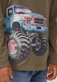 Olivgrünes Langarmshirt mit einer Illustration eines blauen und rostfarbenen Monstertrucks mit großen schwarzen Reifen und pinken Akzenten.