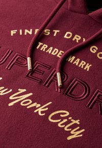 Sudadera burdeos con texto bordado en oro. Presenta cordones con puntas metálicas, resaltando los detalles de la marca y un tejido suave y texturizado.