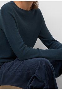 Dunkelgrüner Strickpullover mit Rundhalsausschnitt und langen Ärmeln, kombiniert mit navyblauen gerippten Weitbeinhosen, vor einem neutralen Hintergrund.