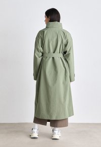 Cappotto trench lungo di colore verde chiaro con vita regolabile, colletto alto e tasche laterali, realizzato in un tessuto liscio. Indossato sopra una gonna marrone.