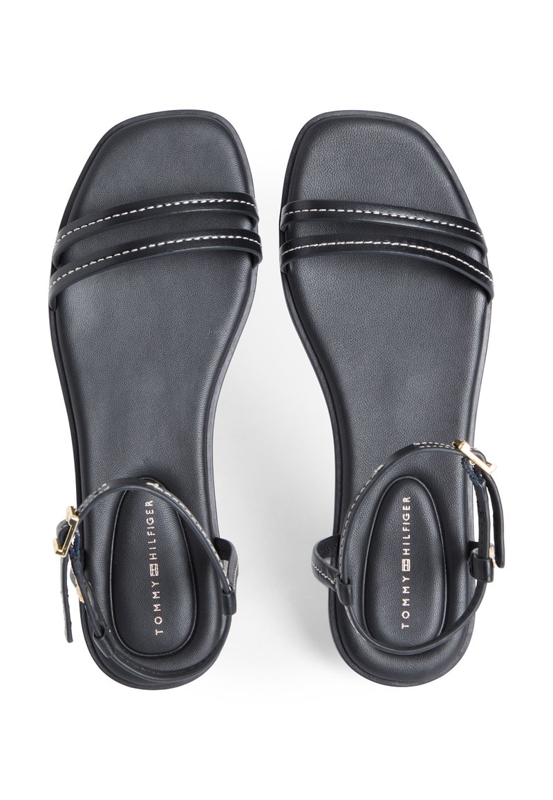 Sandalias planas negras con doble tira frontal, cierres de hebilla en el tobillo y costuras blancas en las tiras, colocadas una al lado de la otra sobre un fondo blanco.