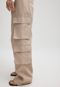 Beige cargobroek met meerdere grote zakken, gemaakt van stevig materiaal. Losse pasvorm met een rechte pijp, gecombineerd met zwarte sandalen.