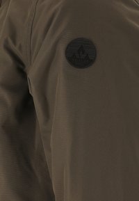 Olivgrön tyg med en slät yta, med en cirkulär svart logopatch märkt "WHISTLER" på övre vänstra sidan.