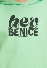 Mintgrüner Hoodie mit großen schwarzen Buchstaben, auf denen steht: "hey BENICE TO YOURSELF." Der Stoff wirkt weich mit einer strukturierten Oberfläche. Verfügt über eine Kapuze mit Kordelzug.