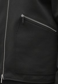 Schwarze Reißverschlussjacke mit einer seitlichen Reißverschlusstasche. Hergestellt aus glattem Stoff, mit silberfarbenen Beschlägen und minimalistischem Design.