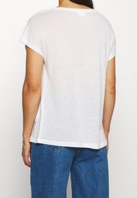 Bílý tričko s krátkým rukávem a volným střihem, jemnou texturou a malou etiketou na zadní straně. V kombinaci s volnými modrými džíny.