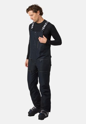 Salopette nera con bretelle regolabili, numerose tasche con zip e vestibilità ampia. Materiale leggero con dettagli di branding minimi.