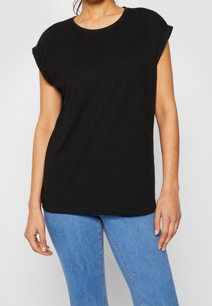 Mujer con camiseta negra de manga corta con puños enrollados y vaqueros azul claro, de pie frente a un fondo gris claro liso.