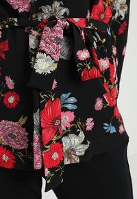 Černá květinová blůza s výraznými červenými, růžovými a modrými květy. Navržena s mašlí v pase a texturovanou tkaninou.