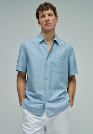 Homme aux cheveux bouclés portant une chemise bleu clair à manches courtes boutonnée et un pantalon blanc, debout les mains dans les poches devant un fond uni.