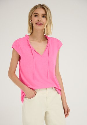 Femme aux cheveux blonds courts portant un chemisier rose vif à manches courtes et un pantalon beige clair, souriante avec une main dans la poche.