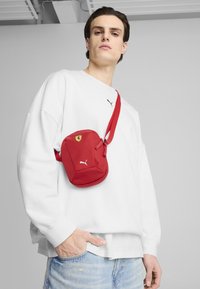 Geantă de umăr roșie Puma cu o suprafață texturată, având un logo galben Ferrari și un emblema albă Puma. Curea roșie ajustabilă.