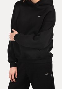 Personne portant un sweat à capuche noir et un pantalon noir assorti avec de petits logos de marque blancs sur la poitrine et la cuisse supérieure.