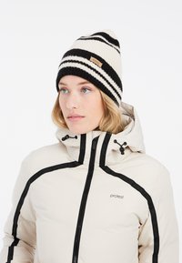 Stribet strikket hue i creme og sort med en spids top og et syet mærke. Båret med en beige pufferjakke med sorte detaljer og lynlåse.