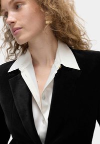 Czarna aksamitna marynarka z białą satynową koszulą pod spodem. Wśród wyróżniających się detali znajdują się stożkowy kołnierz, widoczny guzik oraz fakturowany materiał.