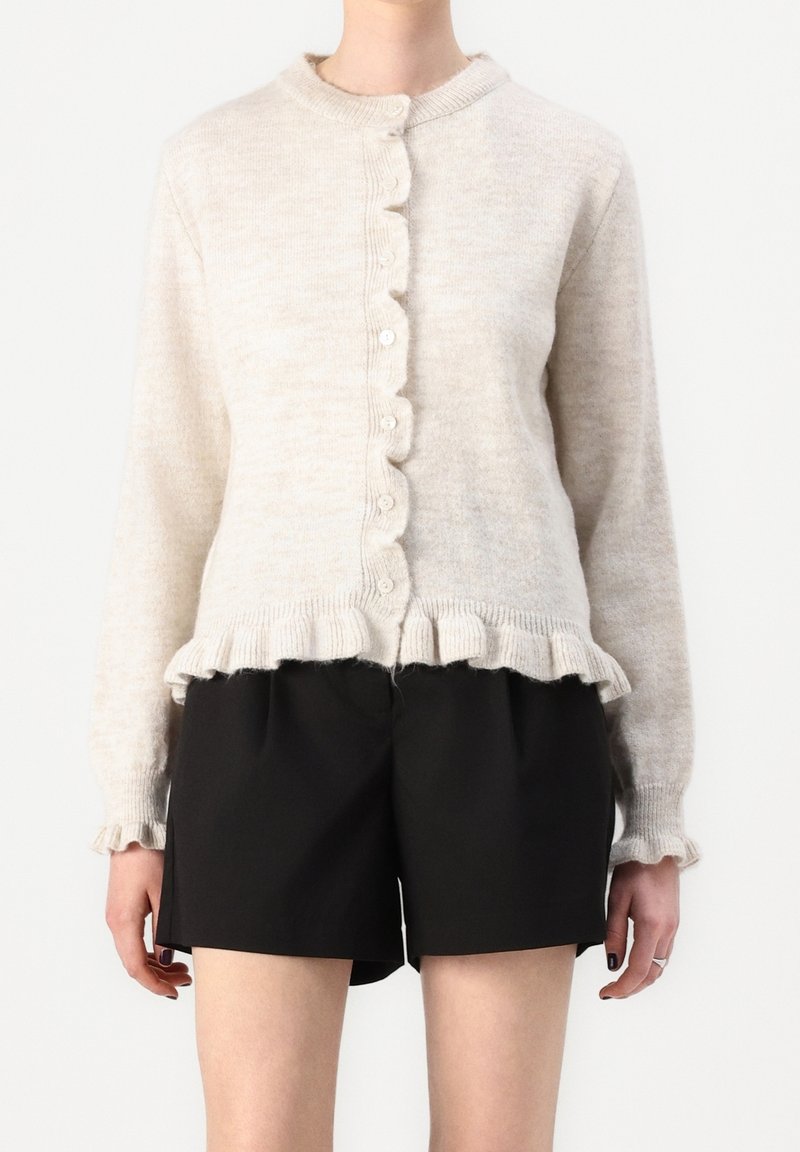 Cárdigan beige claro hecho de una tela suave, con cuello redondo, cierre de botones y dobladillo con volantes. Combinado con pantalones cortos negros.