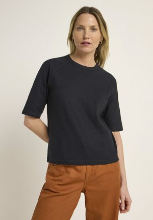 Schwarzes, kurzärmliges T-Shirt mit rundem Halsausschnitt und Raglanärmeln, aus weichem Stoff. Kombiniert mit rostfarbenen Hosen.