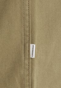 Beżowa tkanina o ukośnej żebrowanej fakturze z widocznym szwem i małą białą metką z napisem „LINDENBERGH” w czerni.