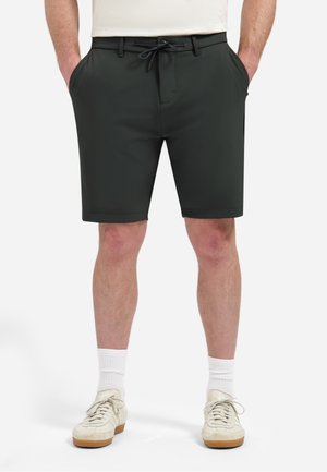 Man met zwarte shorts met trekkoord, witte sokken en crèmekleurige sneakers, staand met handen in de zakken tegen een effen achtergrond.