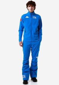 Giacca in pile blu con zip e due tasche laterali, abbinata a pantaloni impermeabili blu coordinati con accenti del logo bianco. Calzature nere.