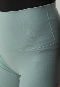 Close-up van een persoon die teal high-waisted Nike leggings draagt met zichtbaar logo en een zwart top.