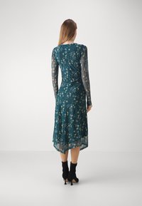 Abito floreale teal con maniche lunghe trasparenti, orlo fluido e corpetto aderente. Stivaletti neri alla caviglia completano l'outfit.