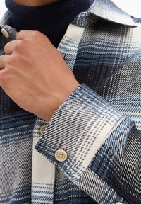 Blue and white plaid flannel shirt, featuring a buttoned cuff and textured fabric, with contrasting navy turtleneck underneath.