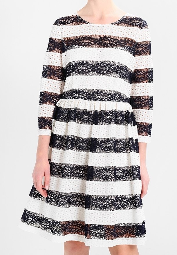 Femme portant une robe mi-longue avec des bandes horizontales en dentelle noire et à œillets blancs, manches trois-quarts et taille froncée.