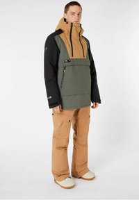 Schneejacke in grün, schwarz und braun mit hohem Kragen und Reißverschlüssen; kombiniert mit hellbraunen Schneehosen. Die Schuhe sind weiß mit braunen Sohlen.