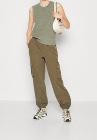 Grünes ärmelloses Stricktop, olivgrüne Cargo-Hosen mit Seitentaschen und beigen Sneakers mit schwarzen Akzenten. Weiße Umhängetasche mit Träger.