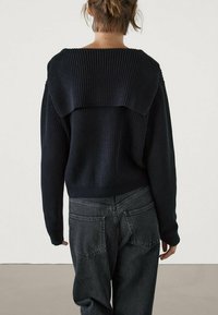 Granatowy sweterek dziany z szerokim kołnierzem na plecach, długimi rękawami i luźnym krojem, w połączeniu z ciemnoszarymi spodniami z wysokim stanem.