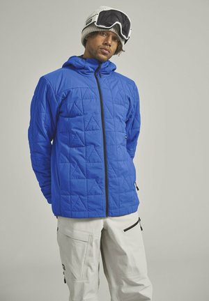 Blue quilted jacket featuring a hood, full-front zipper, and pronounced triangular patterns. Worn with light-colored pants and ski goggles.