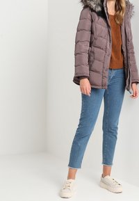 Giacca imbottita di colore marrone chiaro con cappuccio foderato in pelliccia, dotata di chiusura con zip, indossata sopra un maglione marrone e jeans blu, abbinata a sneakers bianche.