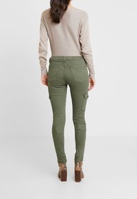 Pantalons cargo skinny vert olive avec poches latérales et poches arrière, associés à un haut à manches longues beige clair et des bottes à talons à motifs.