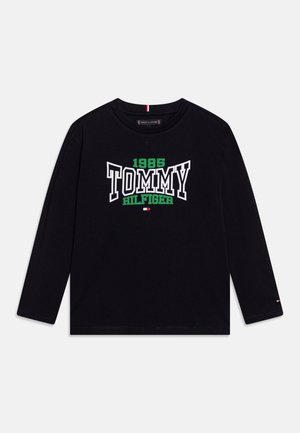 Chemise noire à manches longues avec "1985 Tommy Hilfiger" en lettres blanches et vertes et un petit logo rouge, blanc et bleu sous le texte.