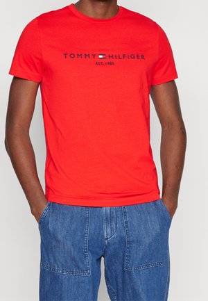 Mężczyzna w jasnoczerwonej koszulce Tommy Hilfiger z logo i niebieskich dżinsach, z rękami w kieszeniach, na jednolitym jasnym tle.