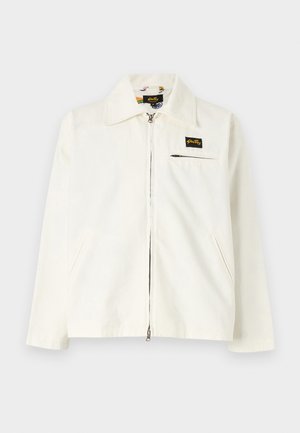White canvas jacket with a collar, front zipper, two side pockets, and a small black logo patch on the left chest.