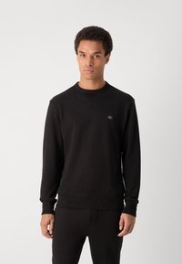Sweatshirt preto feito de um tecido suave, com um decote redondo, punhos canelados e um pequeno patch de logo no peito.