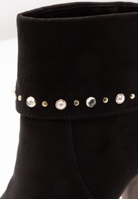 Stivaletto in suede nero con una fila decorativa di borchie in oro e perle lungo il bordo superiore, con una texture liscia e dettagli cuciti.