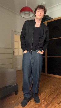 Jaqueta bomber preta com fecho, t-shirt preta, jeans azuis largos, calçado preto, em pé numa sala com chão e móveis de madeira clara.