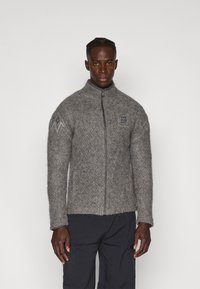 Chaqueta de lana gris con cremallera, con un patrón texturizado y mangas ajustadas, que presenta un alto cuello y un emblema en el lado izquierdo del pecho.