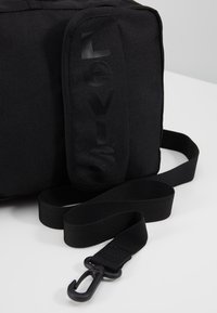 Černá plátěná taška s lesklým logem "LEVIS". Disponuje odnímatelným popruhem a otočným háčkem pro všestrannost. Má strukturovaný, kompaktní design.