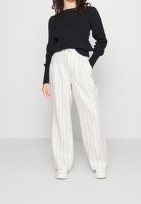 Černý svetr s copánkovým vzorem, dlouhými rukávy a kulatým výstřihem, doplněný bílými širokými kalhotami s pruhy a bílými teniskami.