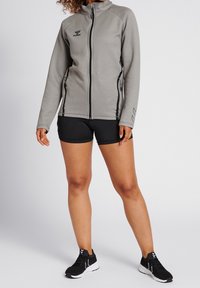 Grå fleecejacka med svarta detaljer, hög krage, framdragkedja och sidofickor. Matchas med svarta shorts och svarta sportskor.