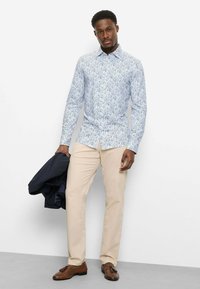 Mann trägt hellblaues Hemd mit Blumenmuster, beige Hose und braune Tasselschuhen, hält einen dunklen Blazer, steht vor weißem Hintergrund.