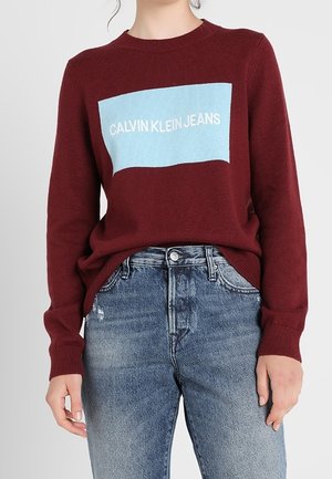 Personne portant un pull bordeaux avec un logo rectangulaire bleu indiquant "Calvin Klein Jeans", associé à un jean bleu taille haute.