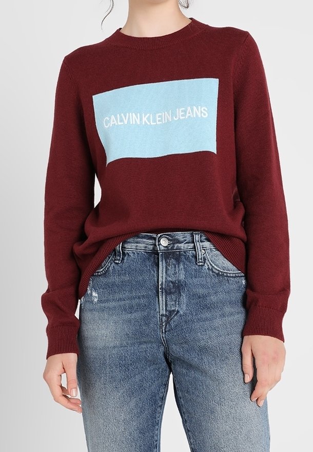 Personne portant un pull bordeaux avec un logo rectangulaire bleu indiquant "Calvin Klein Jeans", associé à un jean bleu taille haute.
