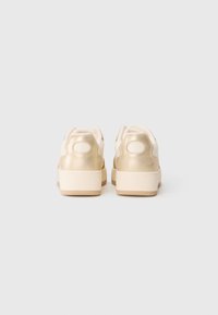 Pair of cream and gold low-top sneakers viewed from the back on a plain white background.