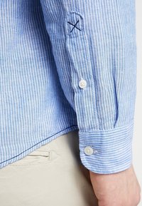 Chemise rayée bleu et blanc avec un poignet boutonné, présentant un détail cousu et une texture douce. Le matériau semble léger et respirant.