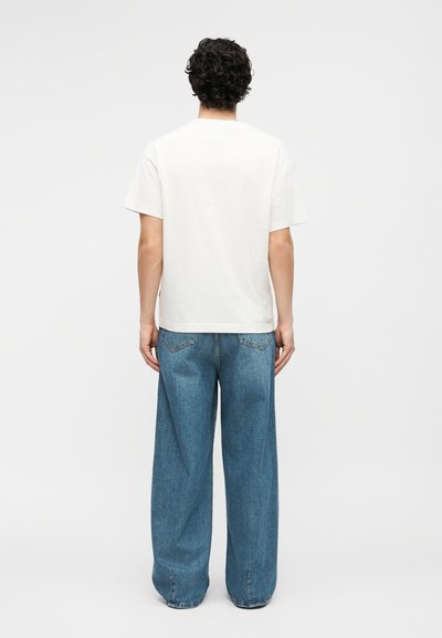 Άνδρας με σγουρά μαλλιά στέκεται με την πλάτη γυρισμένη, φοράει απλό λευκό κοντομάνικο μπλουζάκι και άνετο μπλε τζιν, μπροστά σε λευκό φόντο.